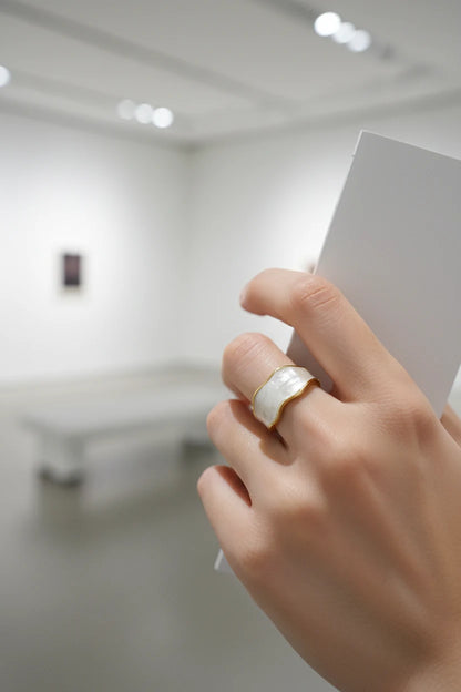 Pearl White Open Wave Ring worn on hand holding a white brochure in a minimalist gallery space, showing the glossy pearl-white enamel band with gold-trim wavy edges and open adjustable design | OriaJewel