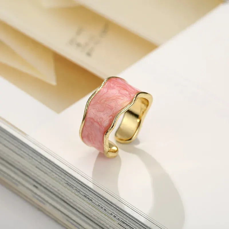 Pink Marble Open Wave Ring macro lifestyle detail resting on the edge of an open book page, warm highlights emphasizing the wavy gold trim and pink marble texture | OriaJewel
