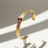 Plum gemstone cuff bracelet displayed upright on a small white stand, with sunlight creating bright highlights on the textured gold band and sparkling off the domed plum stone. | Oria Jewel
