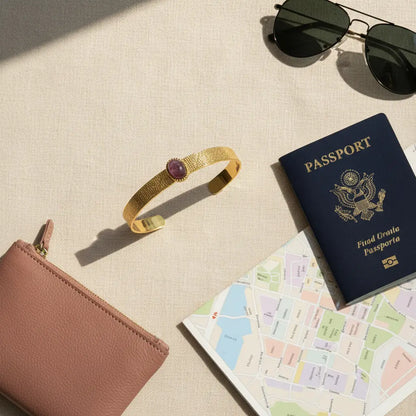Flat-lay of a plum gemstone cuff bracelet on a beige fabric surface, styled with a blush wallet, passport, city map, and sunglasses to suggest a chic travel outfit. | Oria Jewel