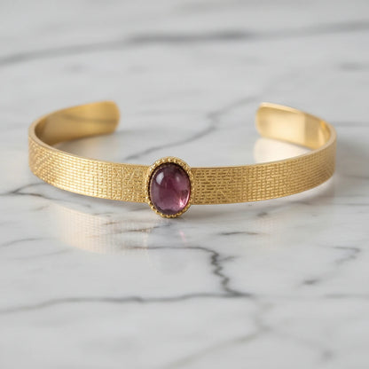 Detailed close-up of the plum gemstone cuff bracelet lying on white marble, highlighting the textured gold band and smooth oval plum stone set in a delicate beaded bezel. | Oria Jewel