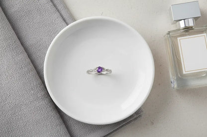 Purple Solitaire Filigree Ring centered on a white ceramic dish beside a folded gray towel and perfume bottle, clean vanity flat lay. | OriaJewel