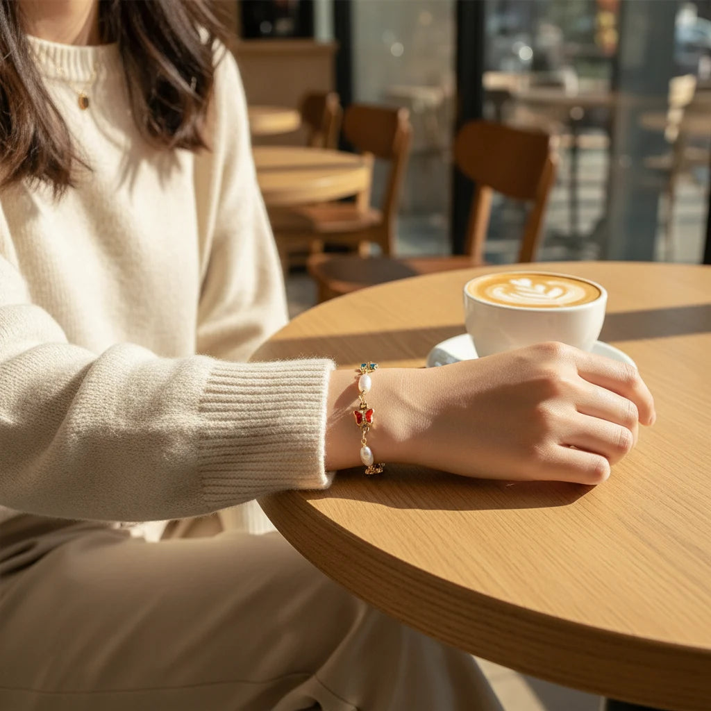 Cozy café scene with a woman in a cream knit sweater resting her arm on a wooden table, showcasing a dainty Rainbow Butterfly Pearl Chain Bracelet with multicolor butterfly charms and freshwater pearls in soft morning light | Oria Jewel