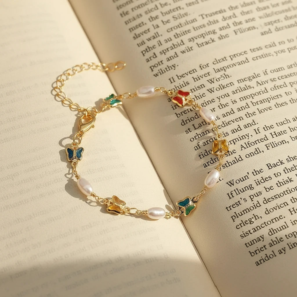 Flat-lay of the Rainbow Butterfly Pearl Chain Bracelet arranged on an open book, the gold chain, pearl accents, and multicolor butterfly charms glowing in warm sunlight for a romantic literary aesthetic | Oria Jewel