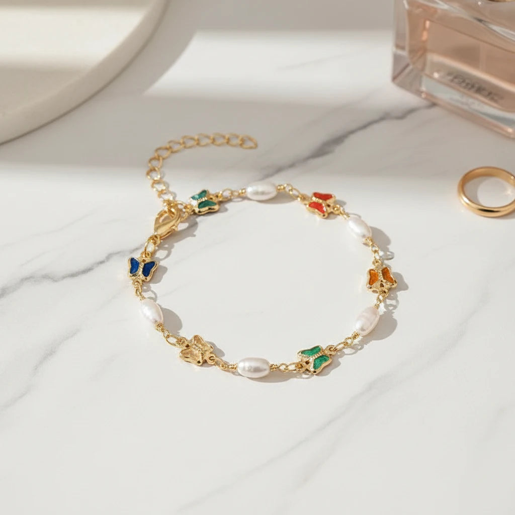Product close-up of the Rainbow Butterfly Pearl Chain Bracelet displayed on white marble, highlighting the glossy enamel butterfly charms, luminous pearls, and adjustable gold chain beside a perfume bottle and ring | Oria Jewel