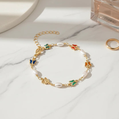 Product close-up of the Rainbow Butterfly Pearl Chain Bracelet displayed on white marble, highlighting the glossy enamel butterfly charms, luminous pearls, and adjustable gold chain beside a perfume bottle and ring | Oria Jewel