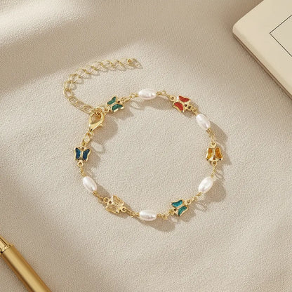 Flat-lay of the Rainbow Butterfly Pearl Chain Bracelet on a soft beige fabric surface, surrounded by stationery items, emphasizing the bracelet’s delicate gold links, alternating pearls, and colorful butterfly charms | Oria Jewel