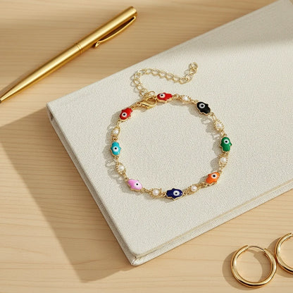 Close-up flat lay of a notebook and gold pen on a wooden desk, centered on the colorful Rainbow Evil Eye Charm Bracelet with tiny charms and pearls | Oria Jewel