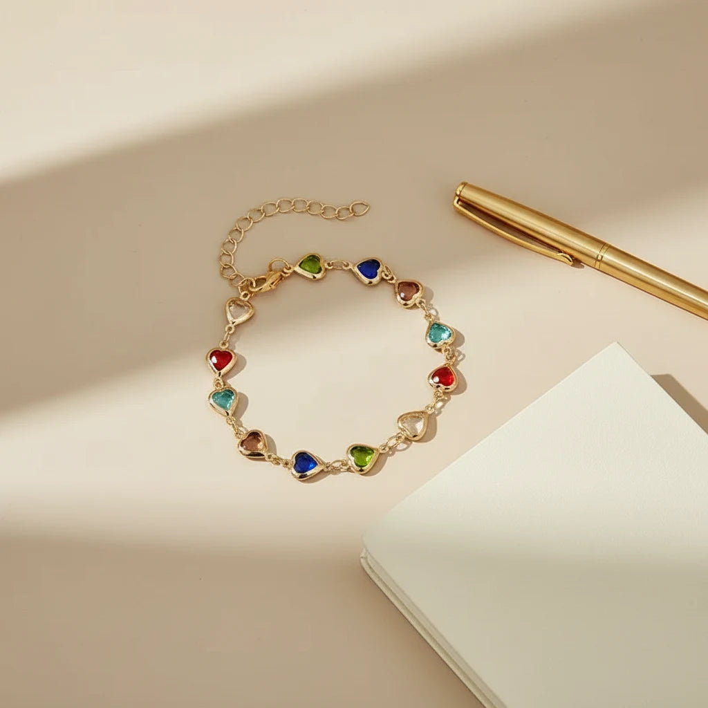 Rainbow Heart Charm Bracelet photographed on a soft beige background with notebook and pen, emphasizing the dainty gold chain and glossy jewel-tone hearts | Oria Jewel