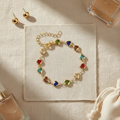 Rainbow Heart Charm Bracelet arranged on a fabric square with perfume bottles and gold stud earrings, highlighting the colorful heart stations as a chic gifting set | Oria Jewel

