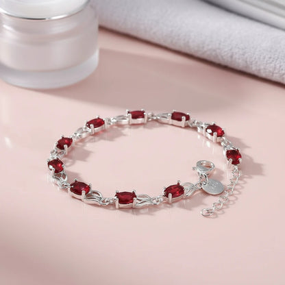 Red Crystal Silver Tennis Bracelet displayed in a loose circle on a glossy blush-pink surface beside a frosted skincare jar and folded towel, highlighting the vibrant oval red stones in delicate silver prong settings | Oria Jewel