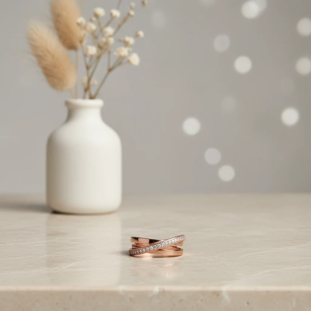 Rose Gold Criss Cross CZ Band Ring resting on a light stone tabletop with a minimal white vase and soft bokeh dots in the background, showcasing the stacked crossover design and sparkling CZ band | OriaJewel
