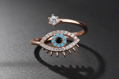 Macro close-up of the Rose Gold Evil Eye Ring on a dark textured surface, showing the sparkling eye outline, turquoise-blue stones around the center, and the small clear stone on the open band | OriaJewel