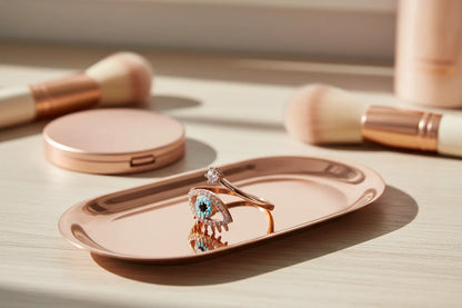 Rose Gold Evil Eye Ring displayed on a rose-gold tray with blurred makeup brushes and a compact in the background, warm morning light reflecting on the metal and stones | OriaJewel