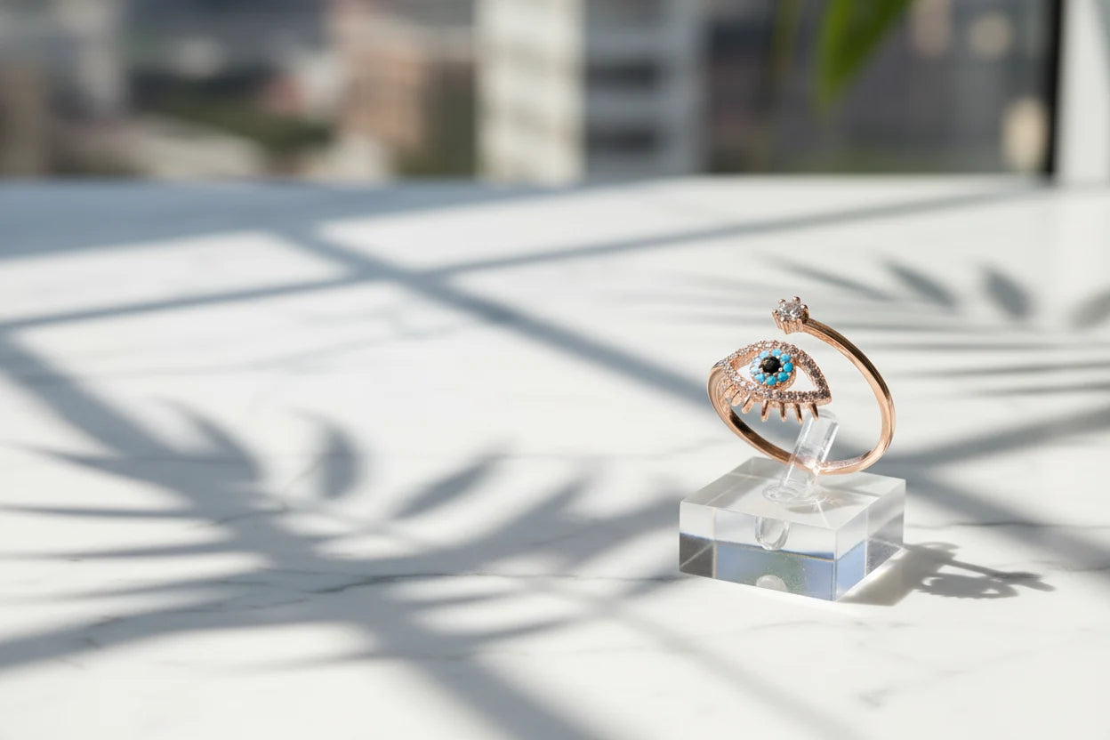 Rose Gold Evil Eye Ring standing on a clear acrylic block on a marble surface, bright window light creating artistic leaf shadows while the evil-eye details stay sharp and centered | OriaJewel