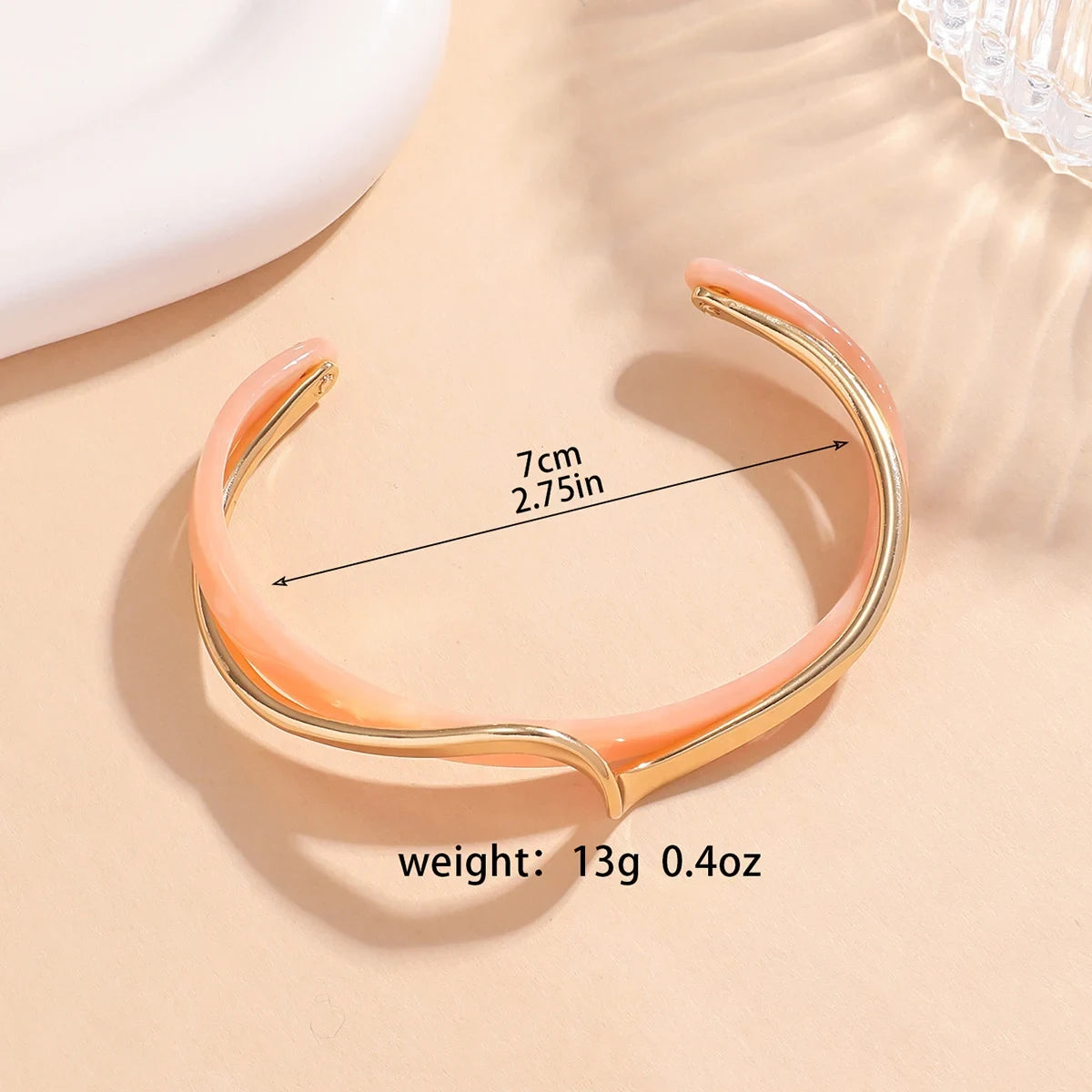 Rosette twist cuff bracelet in blush pink enamel with gold twist detail, shown flat with size 7 cm (2.75 in) and weight 13 g (0.4 oz) text overlay on a soft beige background | Oria Jewel