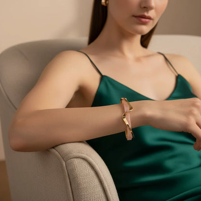 Elegant woman in a deep green satin dress lounging on a cream armchair, showcasing the pink and gold Rosette twist cuff bracelet on her wrist for a chic evening look | Oria Jewel