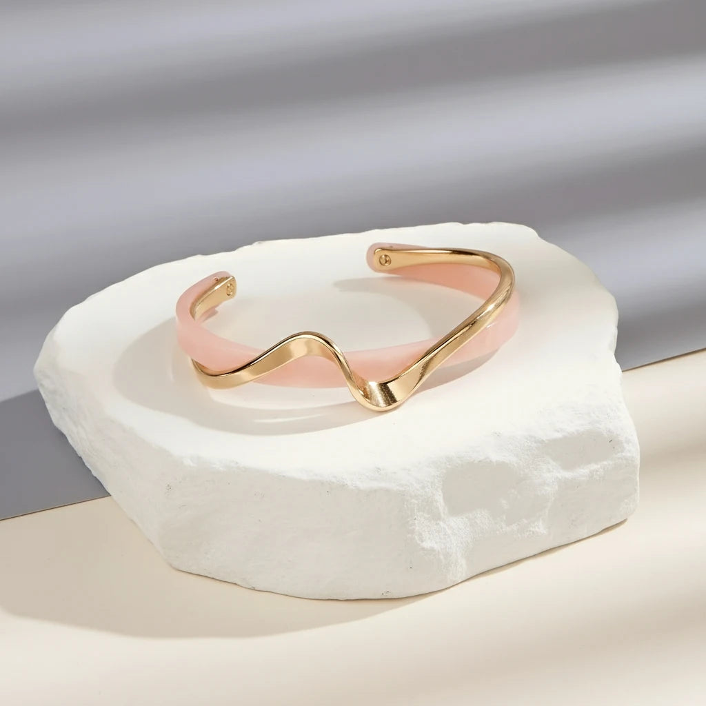 Rosette twist cuff bracelet in pink enamel and gold placed on a textured white stone pedestal, lit by soft studio shadows for a modern jewelry display | Oria Jewel