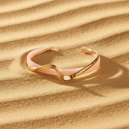 Rosette twist cuff bracelet in pink and gold resting on golden sand, wavy metal design catching warm sunlight and casting soft shadows in the dunes | Oria Jewel