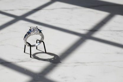 Royal Blue Princess Cut Ring displayed on a small black stand on white marble under window-grid shadows, side/back view highlighting the raised setting, prongs, and blue accent stones | OriaJewel