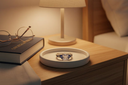 Royal Blue Princess Cut Ring styled on a bedside table in a shallow ceramic tray beside a book, glasses, and lamp, warm ambient lighting highlighting the blue accents and silver setting | OriaJewel
