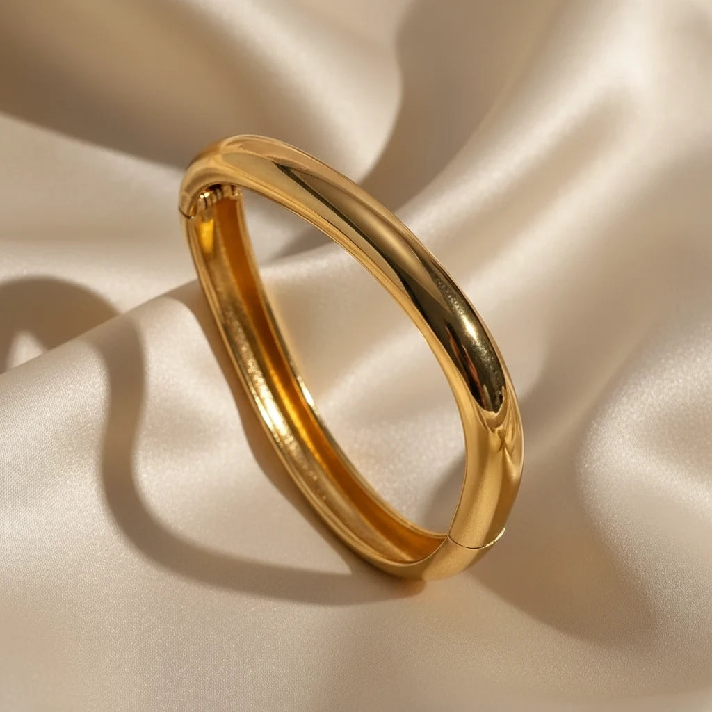 Luxurious close-up of the Sculpted Curve Gold Bangle on soft champagne satin fabric, emphasizing its rounded silhouette and high-shine gold plating for an elegant evening look | Oria Jewel