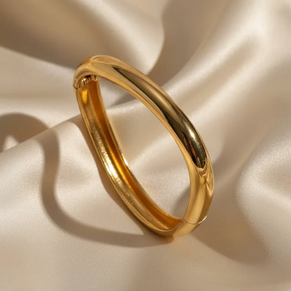 Luxurious close-up of the Sculpted Curve Gold Bangle on soft champagne satin fabric, emphasizing its rounded silhouette and high-shine gold plating for an elegant evening look | Oria Jewel