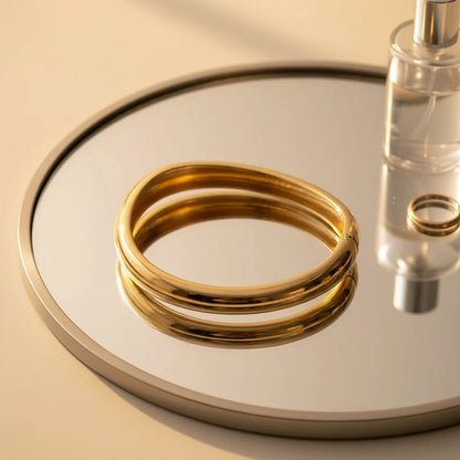 Artistic flat-lay of the Sculpted Curve Gold Bangle doubled over on a round mirror tray, reflecting warm light and beauty essentials to highlight its bold, sculptural profile | Oria Jewel