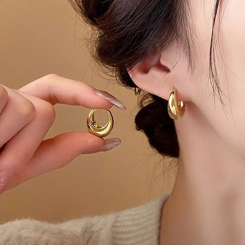 Close-up of shiny chunky gold earrings; one worn, one held by manicured hand | Oria Jewel