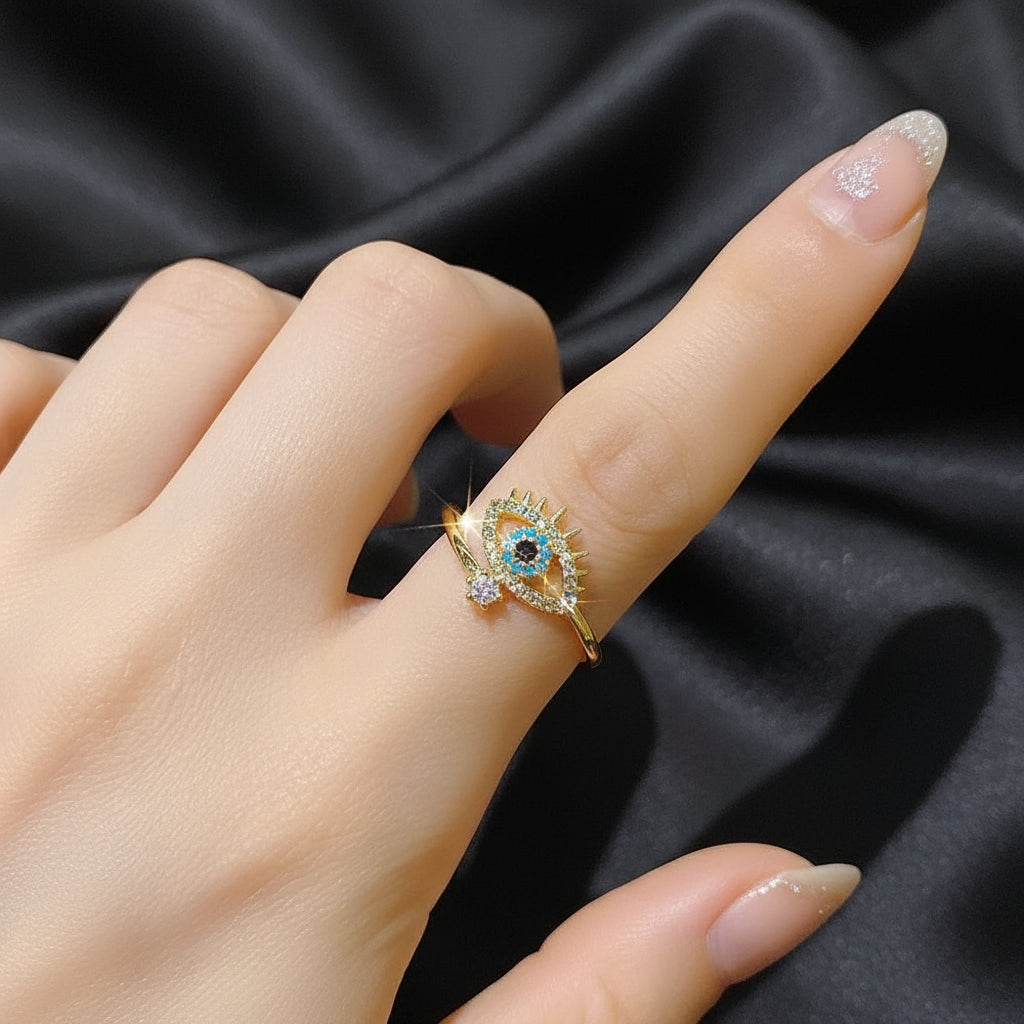 A hand displaying a Blue Crystal Evil Rings with an eye design and gold accents on a dark satin background | Oria Jewel