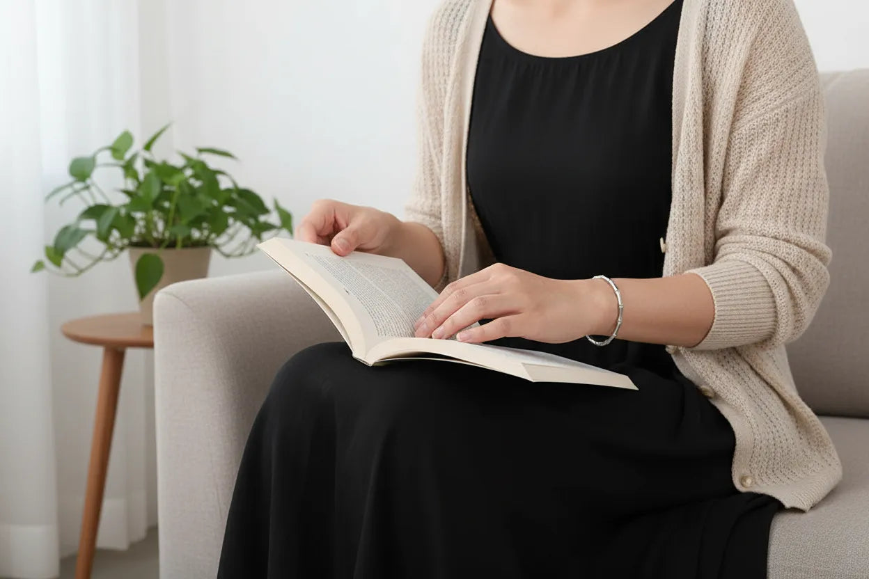 Woman sitting on a sofa reading a book, wearing a delicate Silver Bamboo bangle Bracelet that adds a subtle shine to her casual black dress and cardigan outfit | Oria Jewel
