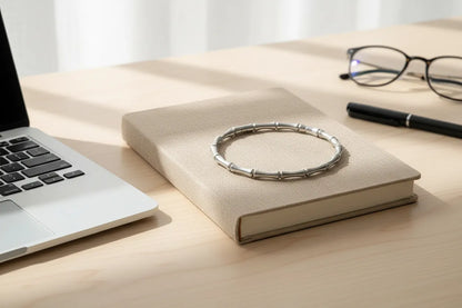 Silver Bamboo bangle Bracelet resting on a beige fabric-covered book beside a laptop, pen, and eyeglasses on a wooden desk, bathed in warm natural window light | Oria Jewel