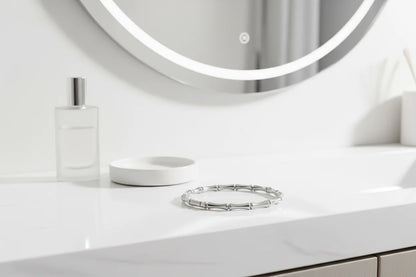 Silver Bamboo bangle Bracelet displayed on a glossy white bathroom counter beneath a round illuminated mirror, with a frosted perfume bottle and dish softly blurred in the background | Oria Jewel
