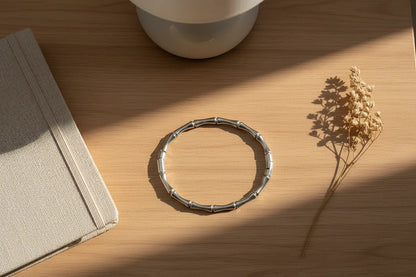 Silver Bamboo bangle Bracelet placed on a wooden surface near a neutral notebook and dried flowers, illuminated by warm striping sunlight for a cozy minimalist workspace feel | Oria Jewel