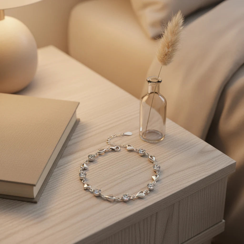 Nightstand flat-lay of the Silver Heart Crystal Tennis Bracelet arranged in a circle beside a closed book and a small glass vase, bathed in soft ambient light that highlights the shimmering crystals and romantic silver hearts | Oria Jewel