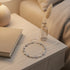 Nightstand flat-lay of the Silver Heart Crystal Tennis Bracelet arranged in a circle beside a closed book and a small glass vase, bathed in soft ambient light that highlights the shimmering crystals and romantic silver hearts | Oria Jewel
