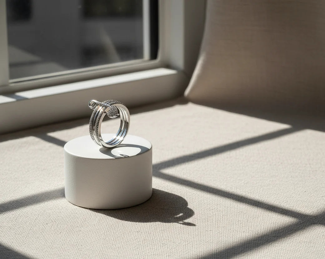 Silver Safety Pin Ring standing on a white cylinder pedestal by a sunny window, crisp grid shadows on a beige fabric surface, clean minimal editorial product shot | OriaJewel