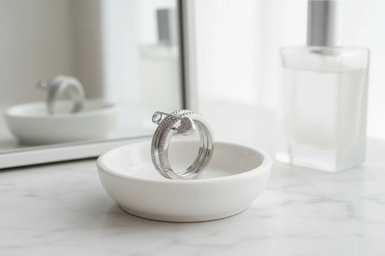 Silver Safety Pin Ring displayed in a white ceramic dish on a marble vanity with soft bathroom lighting, blurred mirror and perfume bottle in the background | OriaJewel