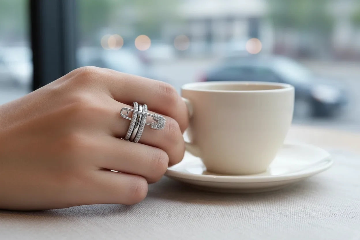 Silver Safety Pin Ring on hand holding a white coffee cup at a café table, close-up focus on the sparkling safety pin and pavé bands with soft city bokeh | OriaJewel