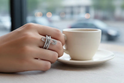 Silver Safety Pin Ring on hand holding a white coffee cup at a café table, close-up focus on the sparkling safety pin and pavé bands with soft city bokeh | OriaJewel