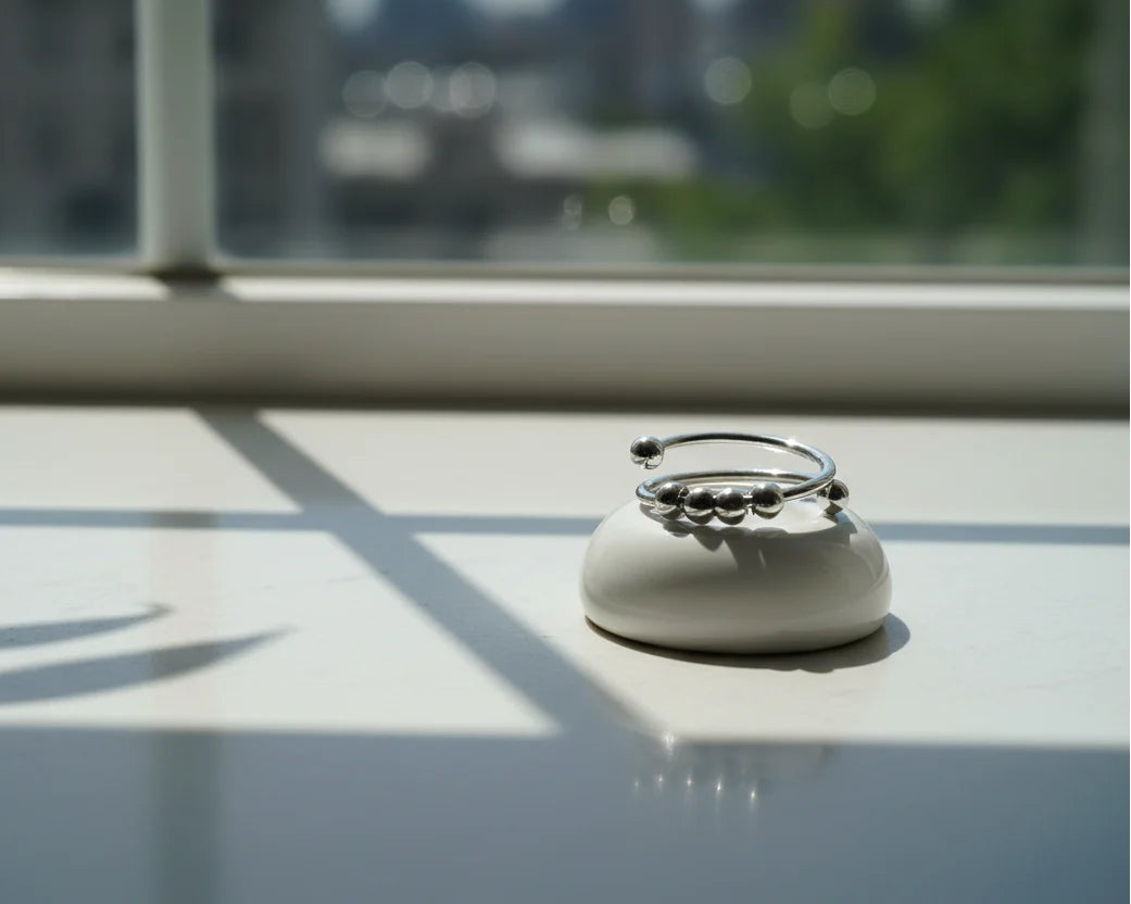 Sunlit windowsill product scene showing the Silver beaded stackable ring displayed on a small ceramic pedestal, bright daylight casting geometric window shadows and a softly blurred city view | OriaJewel