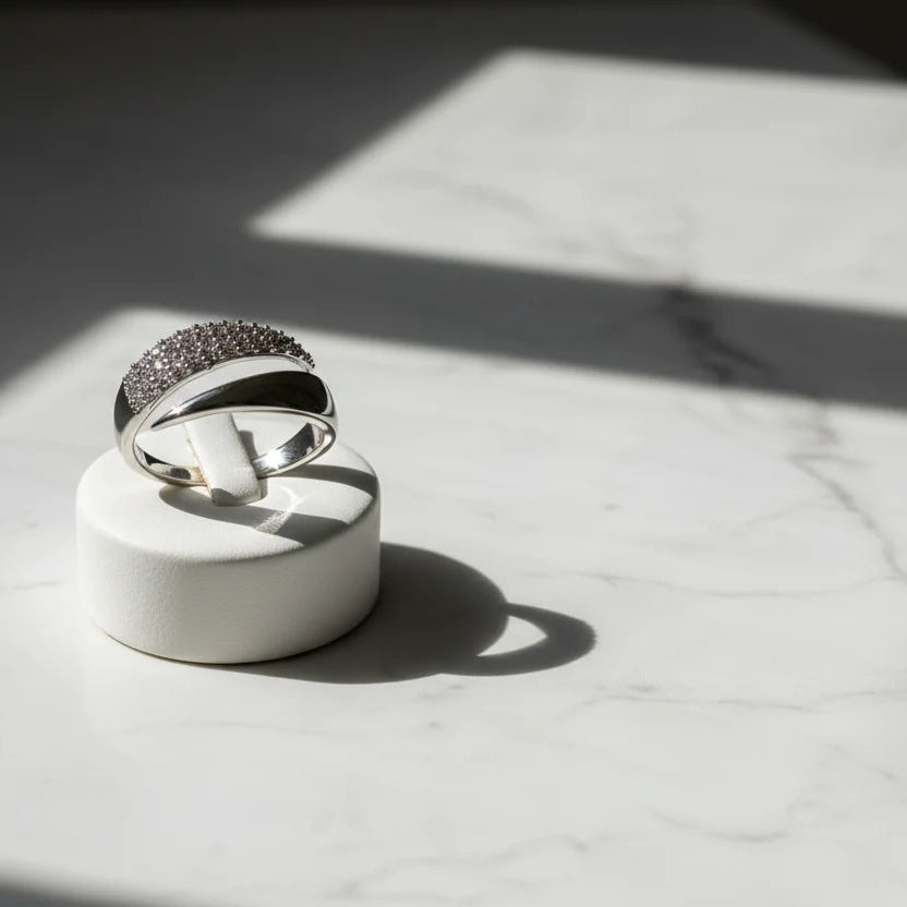 Studio product shot of a Silver bypass ring on a white round stand over marble, with dramatic sunlight and crisp shadows highlighting the pavé curve | OriaJewel