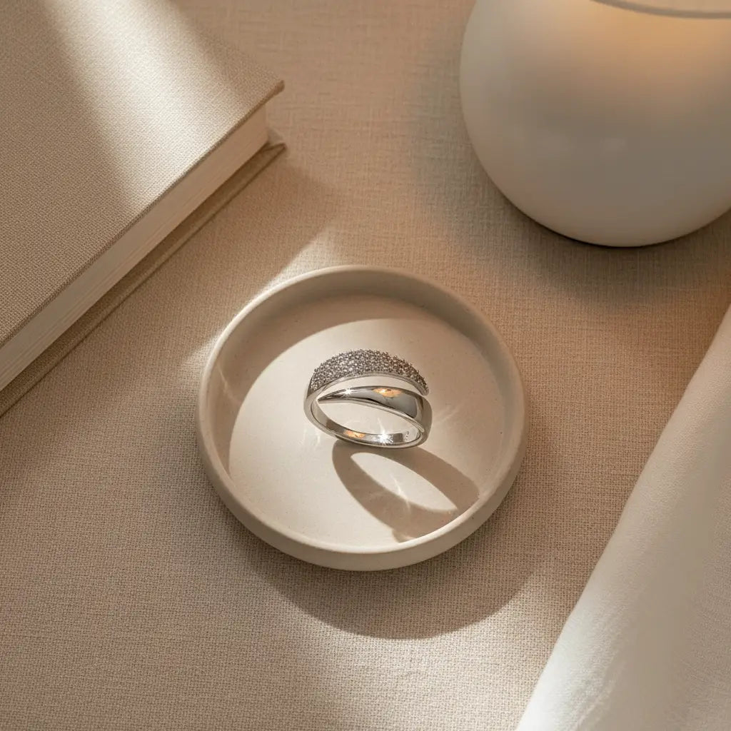 Top-down flat lay of a Silver bypass ring in a ceramic dish on linen, styled with a book and vase, warm sunbeam creating an elegant shadow | OriaJewel