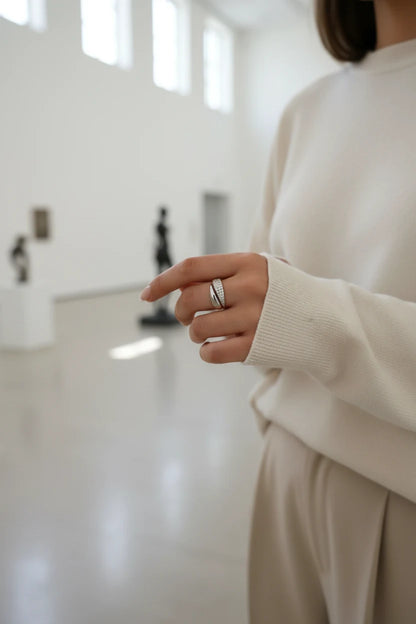 Minimal museum-style scene with a woman in a cream sweater, hand extended to show a Silver bypass ring with pavé shimmer in a bright airy space | OriaJewel