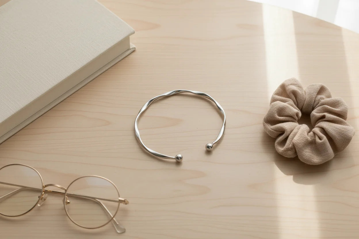Minimal flatlay with Silver twisted cuff bracelet beside round glasses, a beige scrunchie, and a neutral hardcover book on a light wooden desk | Oria Jewel