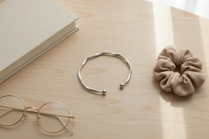 Minimal flatlay with Silver twisted cuff bracelet beside round glasses, a beige scrunchie, and a neutral hardcover book on a light wooden desk | Oria Jewel