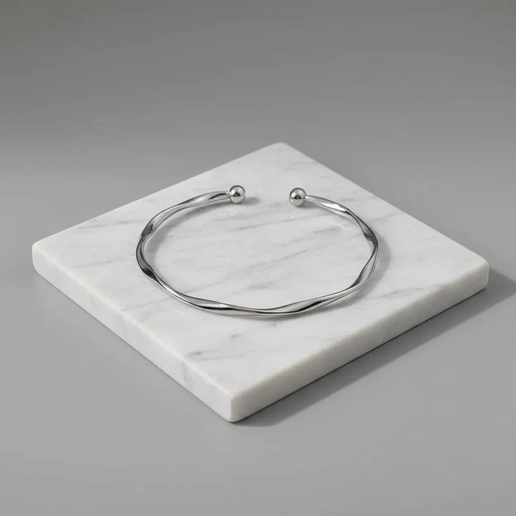 Silver twisted cuff bracelet displayed on a square white marble slab against a soft grey background, emphasizing its smooth curves and polished shine | Oria Jewel