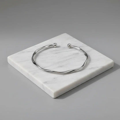 Silver twisted cuff bracelet displayed on a square white marble slab against a soft grey background, emphasizing its smooth curves and polished shine | Oria Jewel
