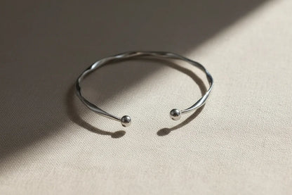Silver twisted cuff bracelet lying open on a taupe fabric surface, sunlight casting gentle shadows that accentuate its twisted design and round ends | Oria Jewel