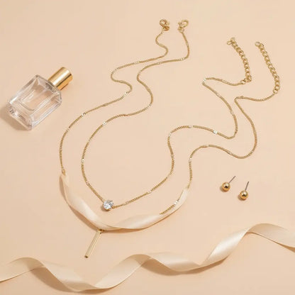 Flat lay of gold Star Layered Necklace arranged with matching gold stud earrings, ribbon, and perfume bottle on peach background | Oria Jewel